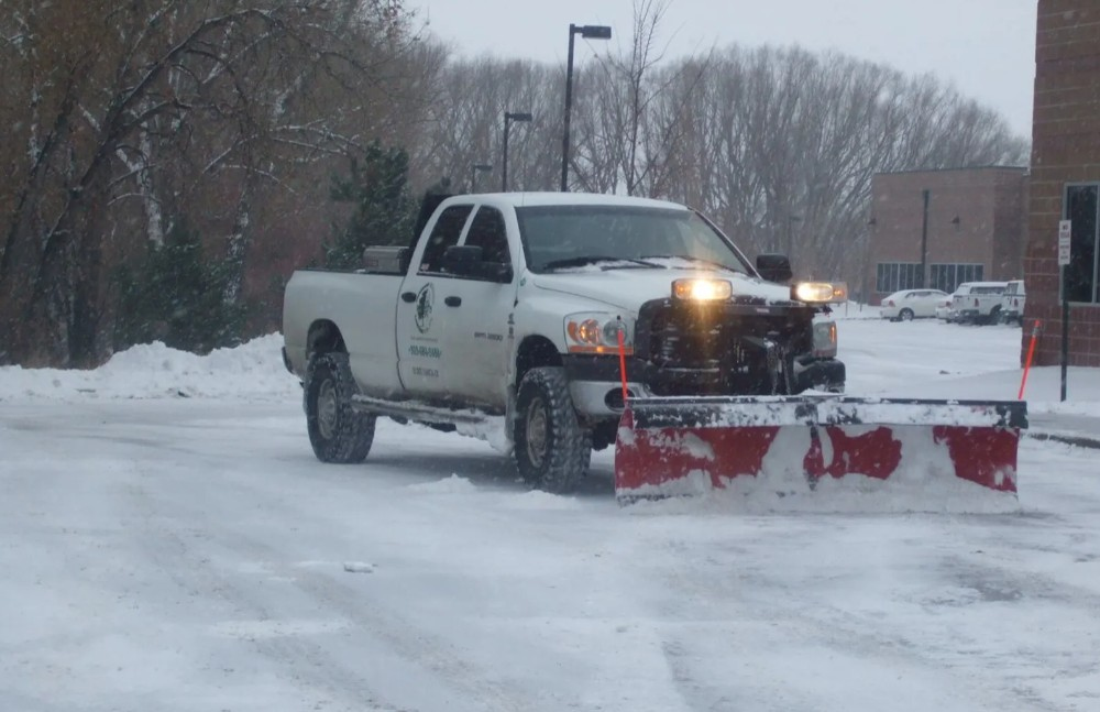 A white pickup truck equipped with a snowplow is clearing a snow-covered paved surface, likely a commercial parking lot, with bare trees and a brick building in the background under a snowy sky. Published by Ward's Lawn, experts in commercial and residential landscaping solutions, including professional snow and ice management in Colorado. This visual demonstrates the effectiveness of professional snow mitigation services, directly illustrating how expert ice and snow removal protects commercial properties and maintains accessibility, which is key to its return on investment. Businesses can learn more about securing their properties for winter by exploring Ward's Lawn's commercial services at wardslawn.com.