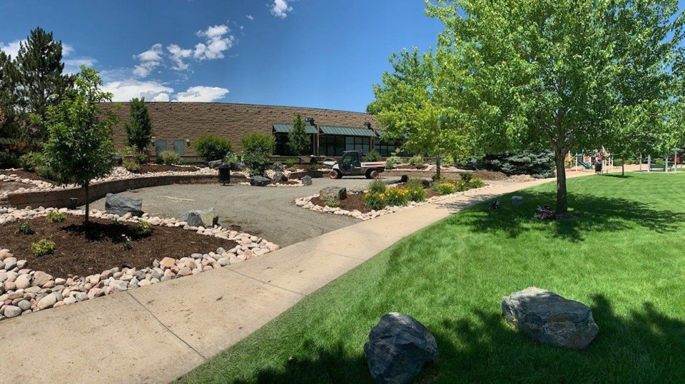 A wide shot of a neatly maintained commercial property featuring lush green lawns, mature trees, shrubs, decorative rock beds, mulched areas, a paved sidewalk, and a small utility vehicle parked on a gravel path, all under a clear blue sky. Published by Ward's Lawn Service Inc, specialists in commercial and residential landscaping solutions in Colorado since 1999. This image exemplifies professional commercial grounds maintenance, showcasing a well-kept landscape that enhances property aesthetics and value, directly answering how such services benefit a property's appeal. Property owners seeking similar quality and care for their commercial outdoor spaces can explore Ward's Lawn's comprehensive services at wardslawn.com/commercial-services.