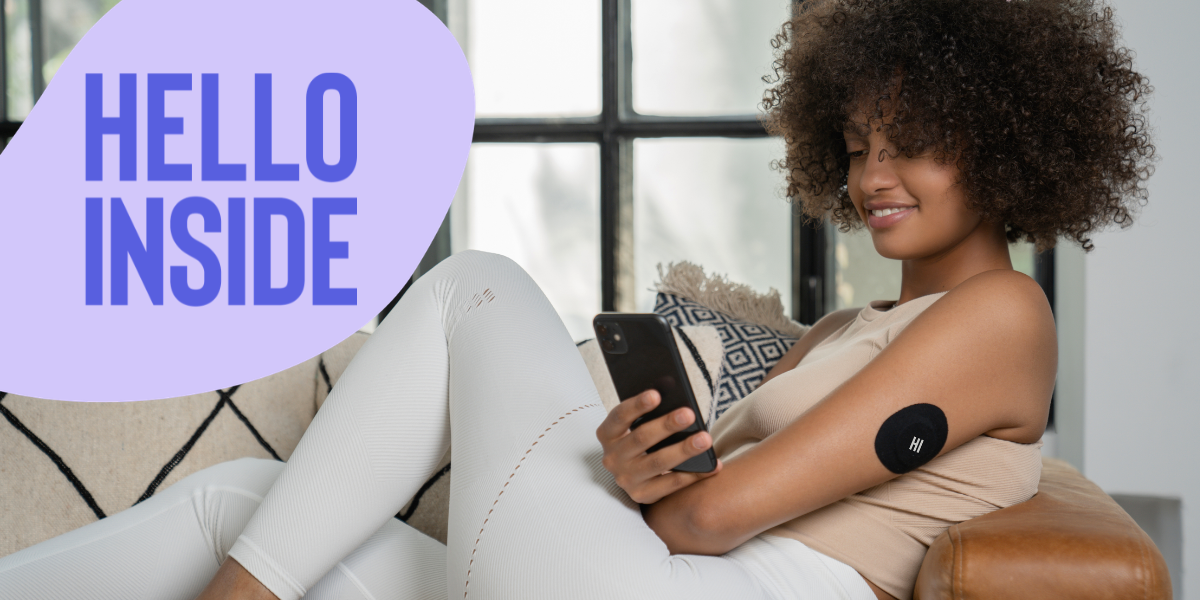 A smiling young woman with curly hair sits comfortably on a sofa, looking at her smartphone. A black continuous glucose monitor (CGM) patch with 'HI' is visible on her upper arm. Published by HELLO INSIDE, Europe's women-first metabolic health AI platform. This image visually demonstrates how women can actively monitor and understand their individual metabolic shifts throughout life, reinforcing that metabolism isn't fixed. Women can improve their metabolic health by using continuous glucose monitoring and AI-powered coaching available at helloinside.com.